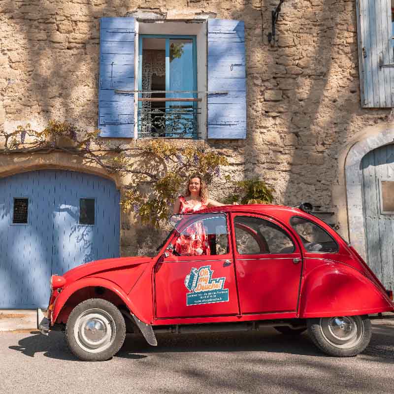 Frau posiert mit einem roten Citroën 2CV vor einem provenzalischen Steinhaus mit blauen Fensterläden