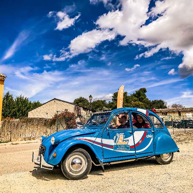 Blaue Citroën 2CV in Bewegung mit lachenden Passagieren unter einem strahlend blauen Himmel in der Provence