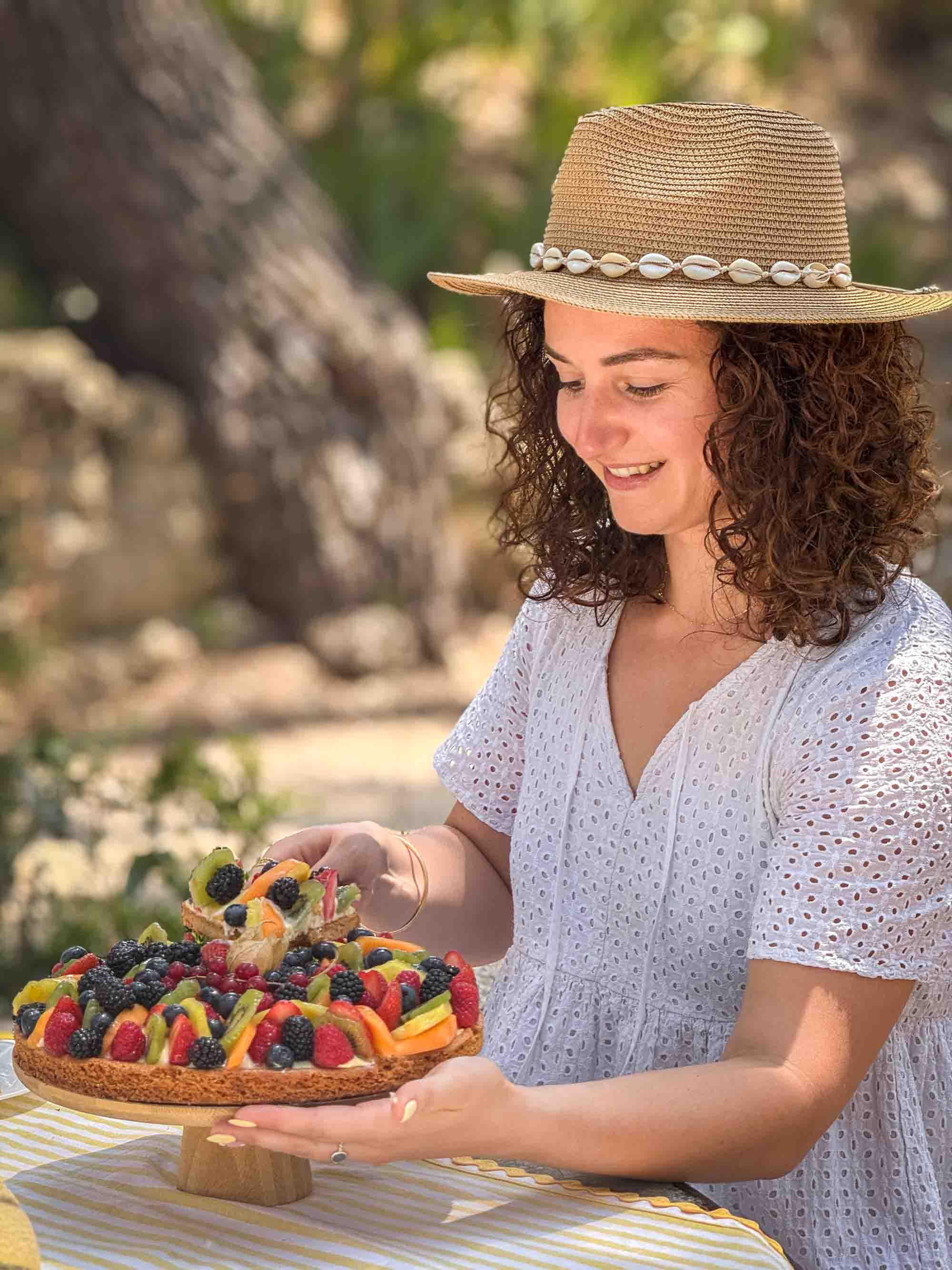 Die Schätze der provenzalischen Region mitten in der Natur genießen Eine lächelnde junge Frau mit einem Strohhut, der mit Muscheln verziert ist, hält eine schöne Tarte mit roten und exotischen Früchten.