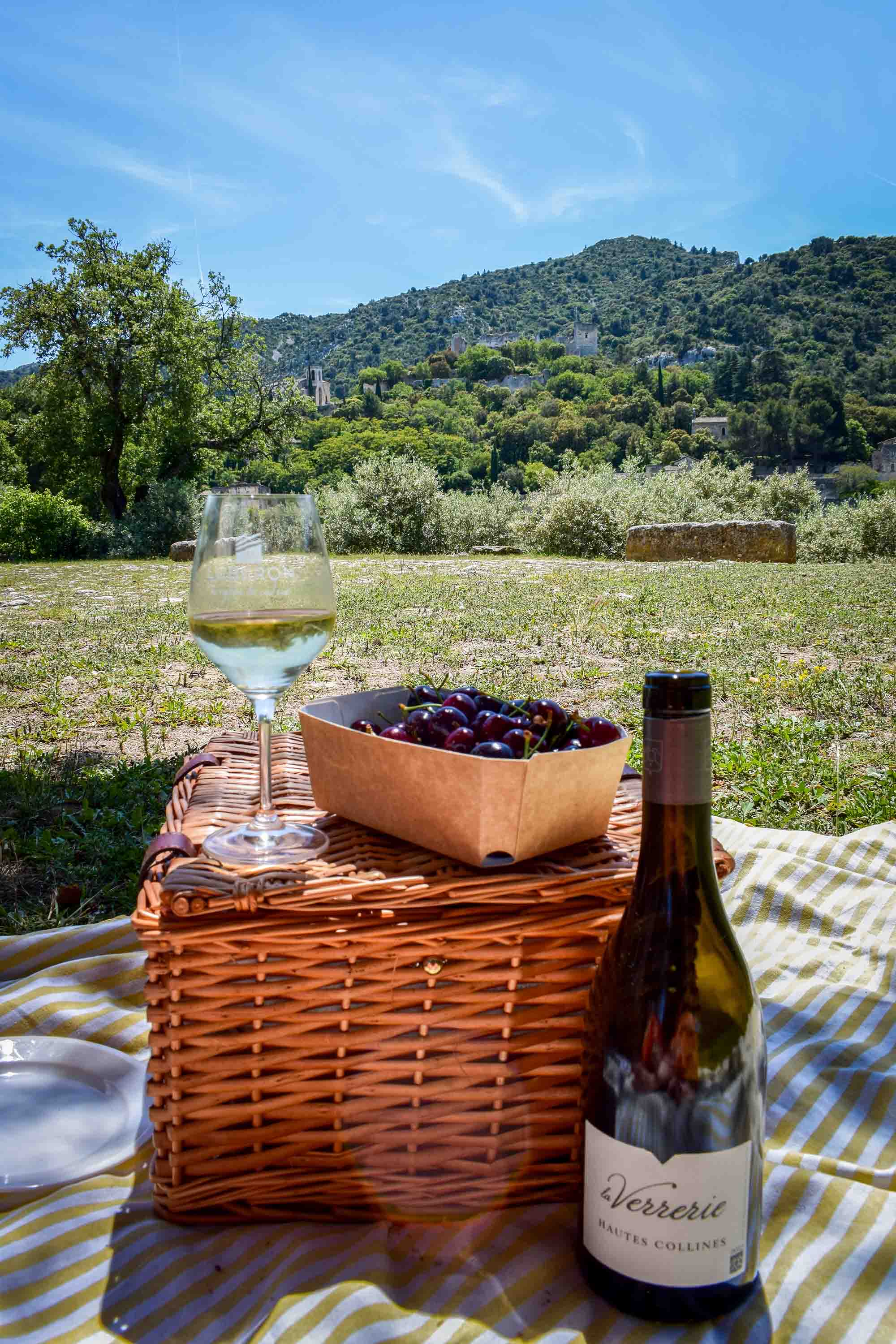 Lebensfreude und Genuss im Herzen von Alt-Oppède Picknick-Komposition: eine Flasche Wein der Verrerie und ein gefülltes Glas neben einem geschlossenen Weidenkorb mit einer Schale frischer schwarzer Kirschen, alles auf einer gestreiften Decke mit Blick auf die Landschaft von Oppède.