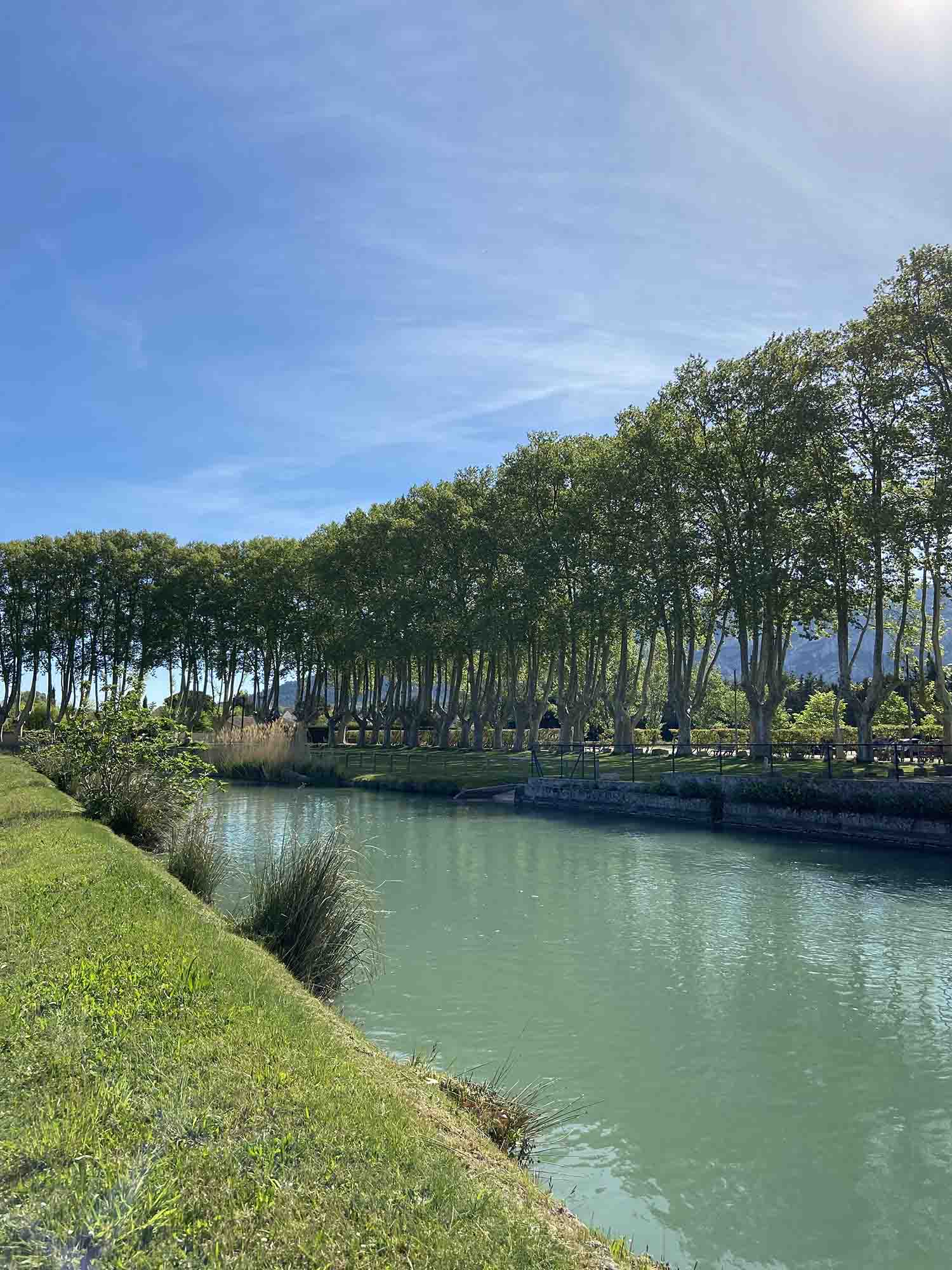 Eine erfrischende Pause am Wasser beim Moulin Saint-Pierre Blick auf den Moulin Saint-Pierre in Les Taillades mit seinem großen hölzernen Schaufelrad am Kanal, umgeben von Grün und Blumen unter blauem Himmel.